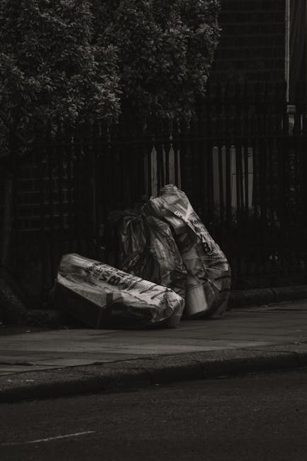 Two waste collection workers are pictured at an urban roadside, operating a large, orange skip loader truck during dusk or evening. The worker in the foreground has his back turned, wearing a turquois
